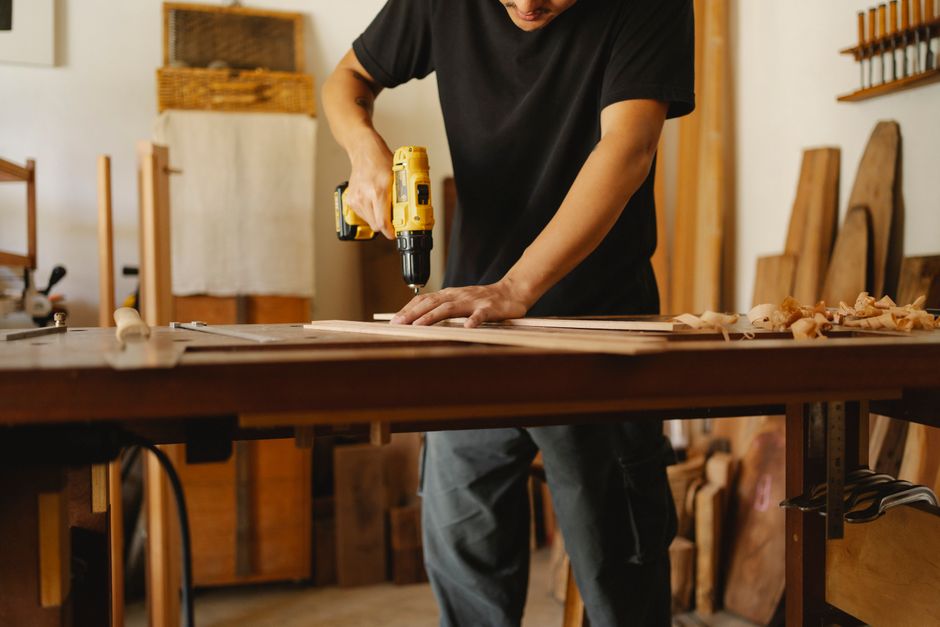 Crop anonymous man in casual clothes standing near workbench and drilling while working in joinery